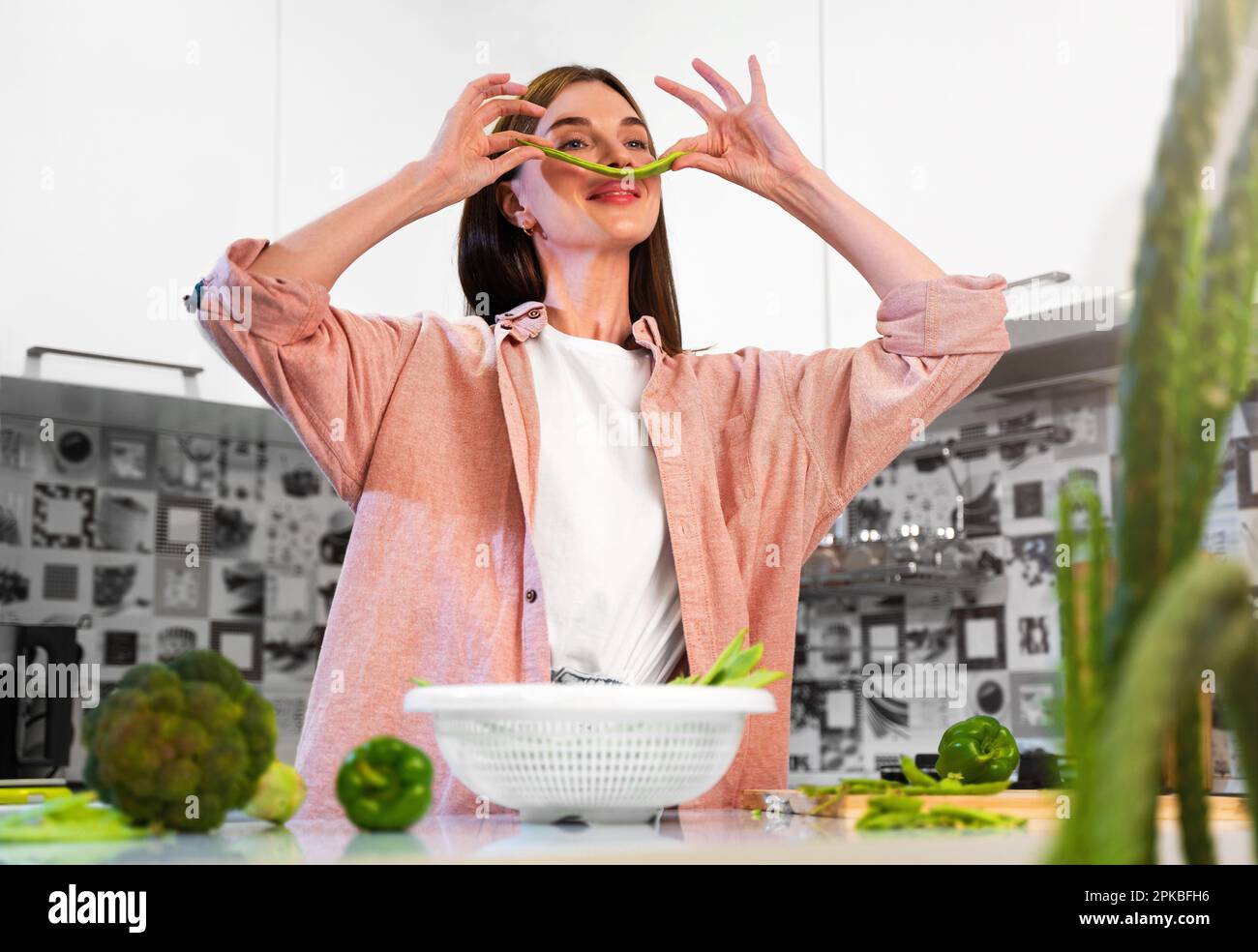 Young beautiful woman making mustache from green bean on face at home kitchen and smiles. The concept of a healthy nutrition and vegan lifestyle.Healt Stock Photo