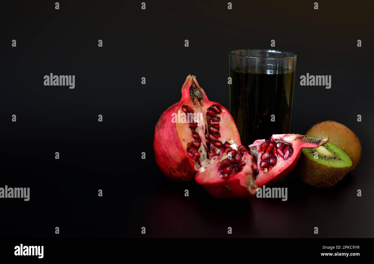 A glass of a mixture of freshly squeezed exotic fruits on a black background, next to a broken pomegranate fruit and pieces of ripe kiwi. Close-up. Stock Photo