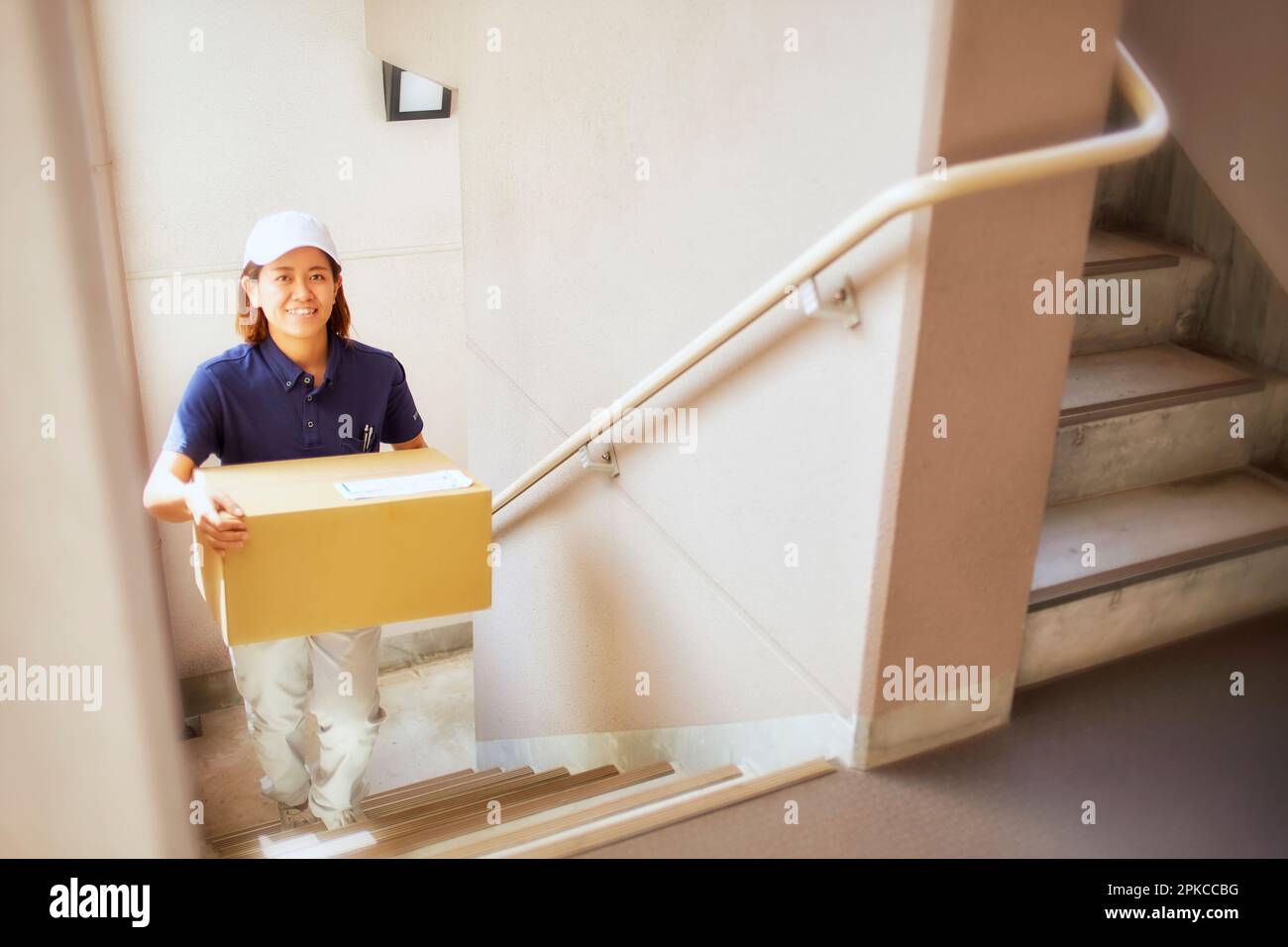 Woman in work clothes carrying a cardboard box up the stairs of an apartment building Stock Photo