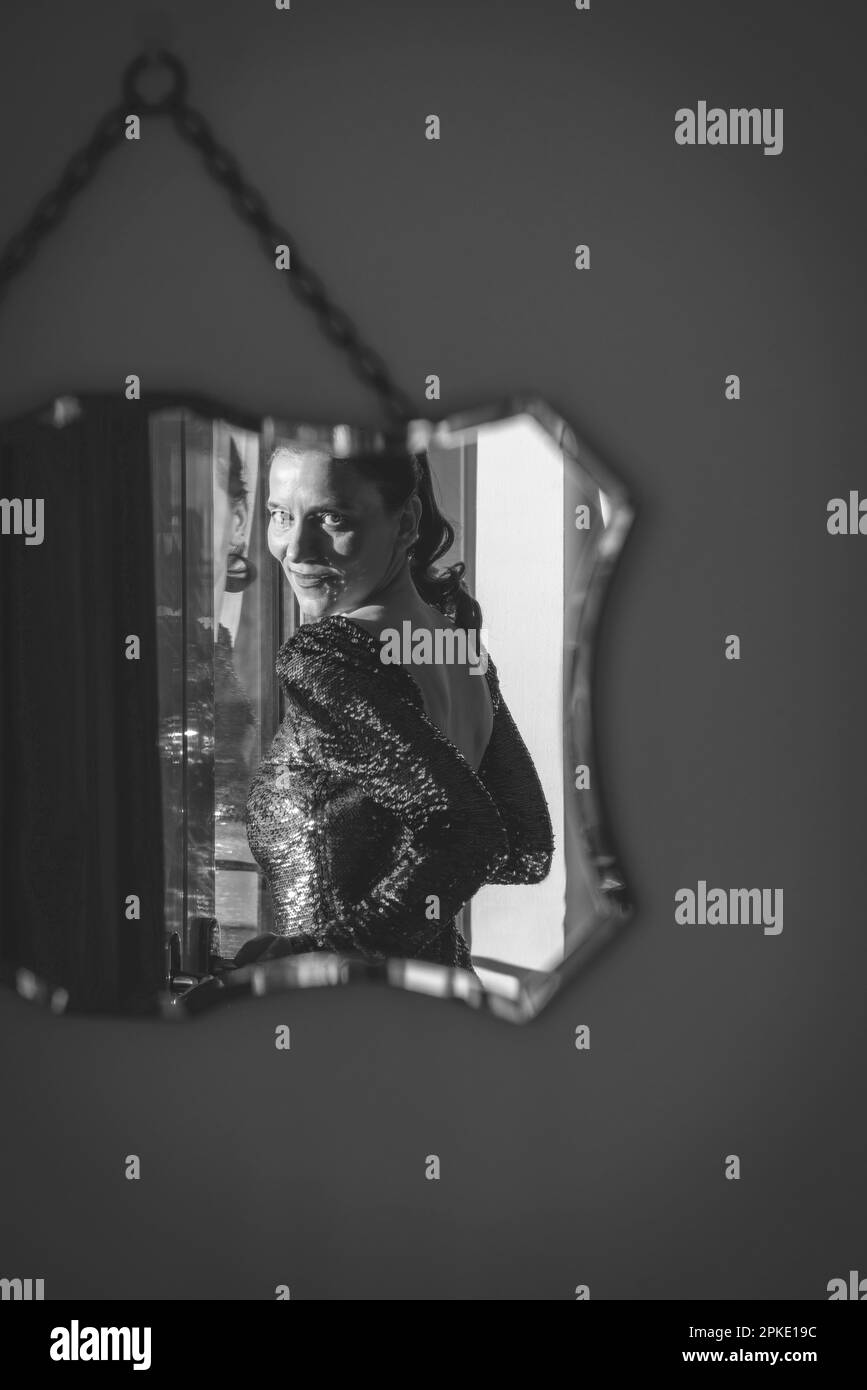 Reflection in a wall mounted mirror of a woman smiling and wearing a sequined party dress - black and white image Stock Photo