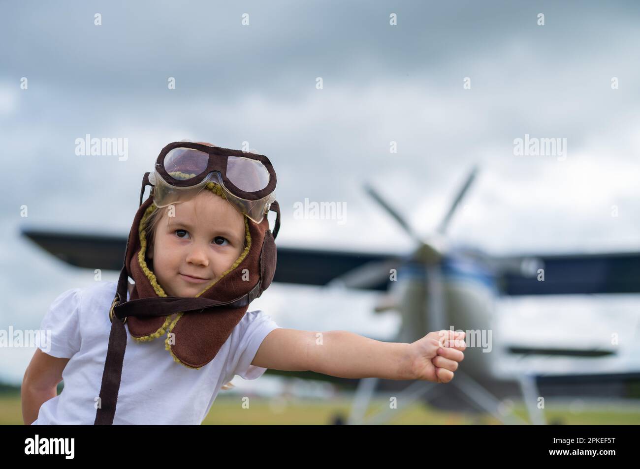 A cute little girl dressed in a cap and glasses of a pilot on the background of an airplane. The child dreams of becoming a pilot. Stock Photo
