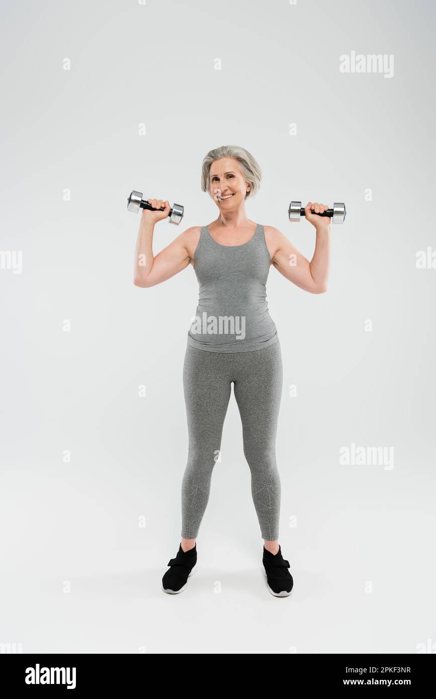 full length of happy senior woman in tight sportswear exercising with dumbbells on grey,stock image Stock Photo