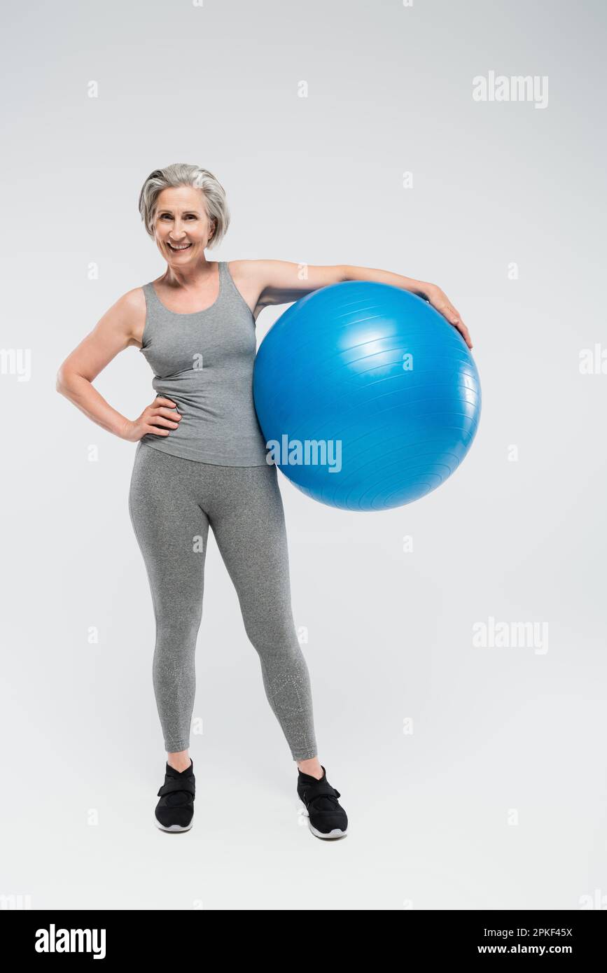 full length of happy senior woman in sportswear holding  fitness ball on grey,stock image Stock Photo