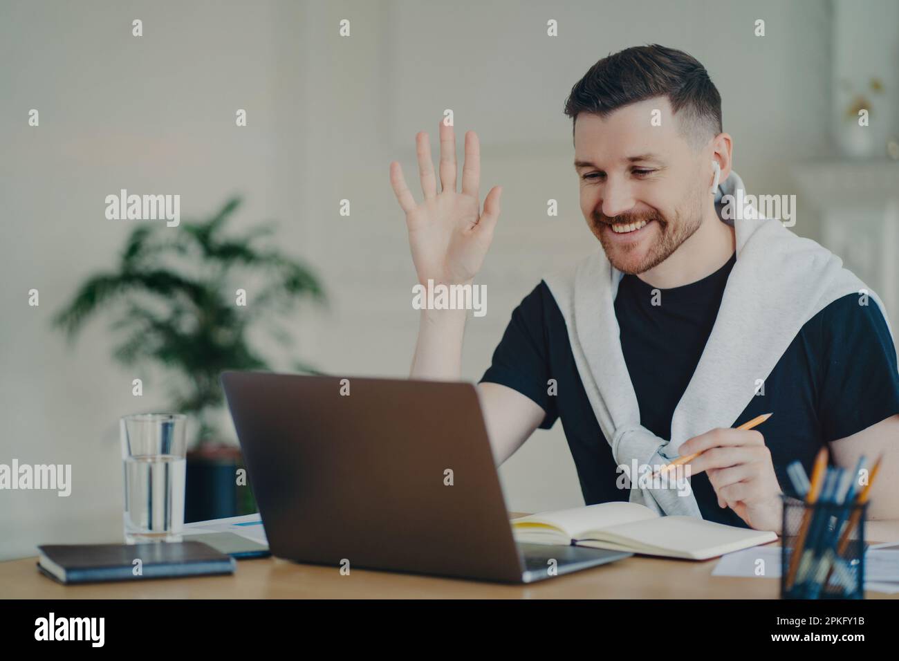 Cheerful bearded man has video conference online waves palm in laptop computer uses wireless earphones poses at coworking space dressed casually write Stock Photo