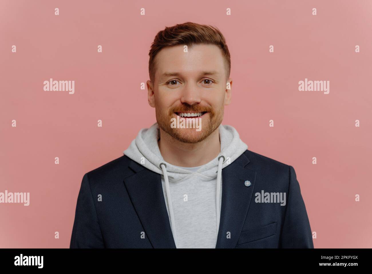 Portrait of positive attractive red haired guy wearing hoodie and jacket smiling at camera, young good looking cheerful man with beard posing in styli Stock Photo