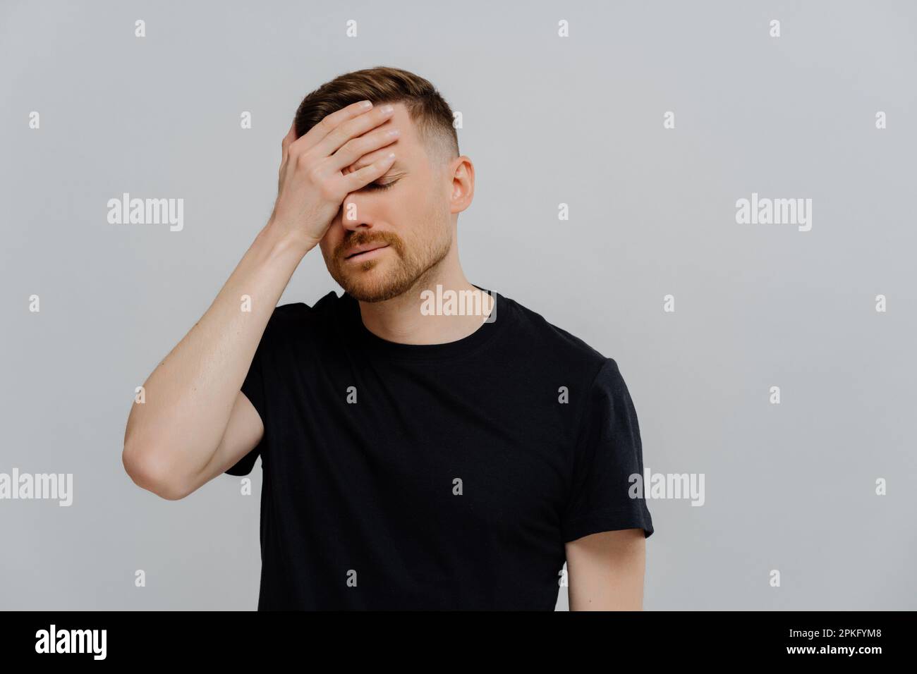 Young tired bearded man in black tshirt with closed eyes feeling sleepy, doing facepalm gesture while posing on pastel rose background, putting hand o Stock Photo