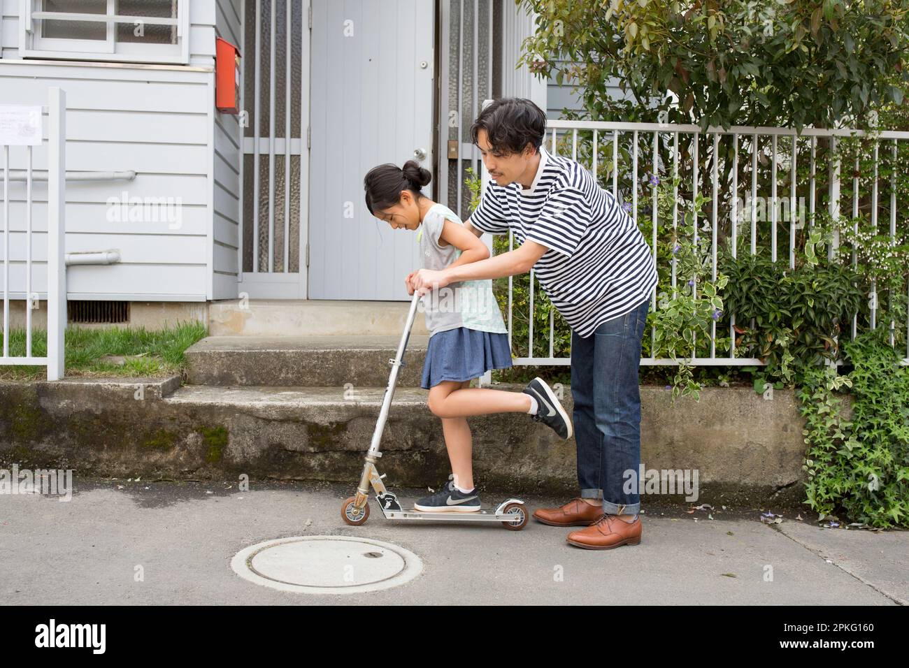 Father teaching his daughter how to ride a kickboard in front of their house Stock Photo