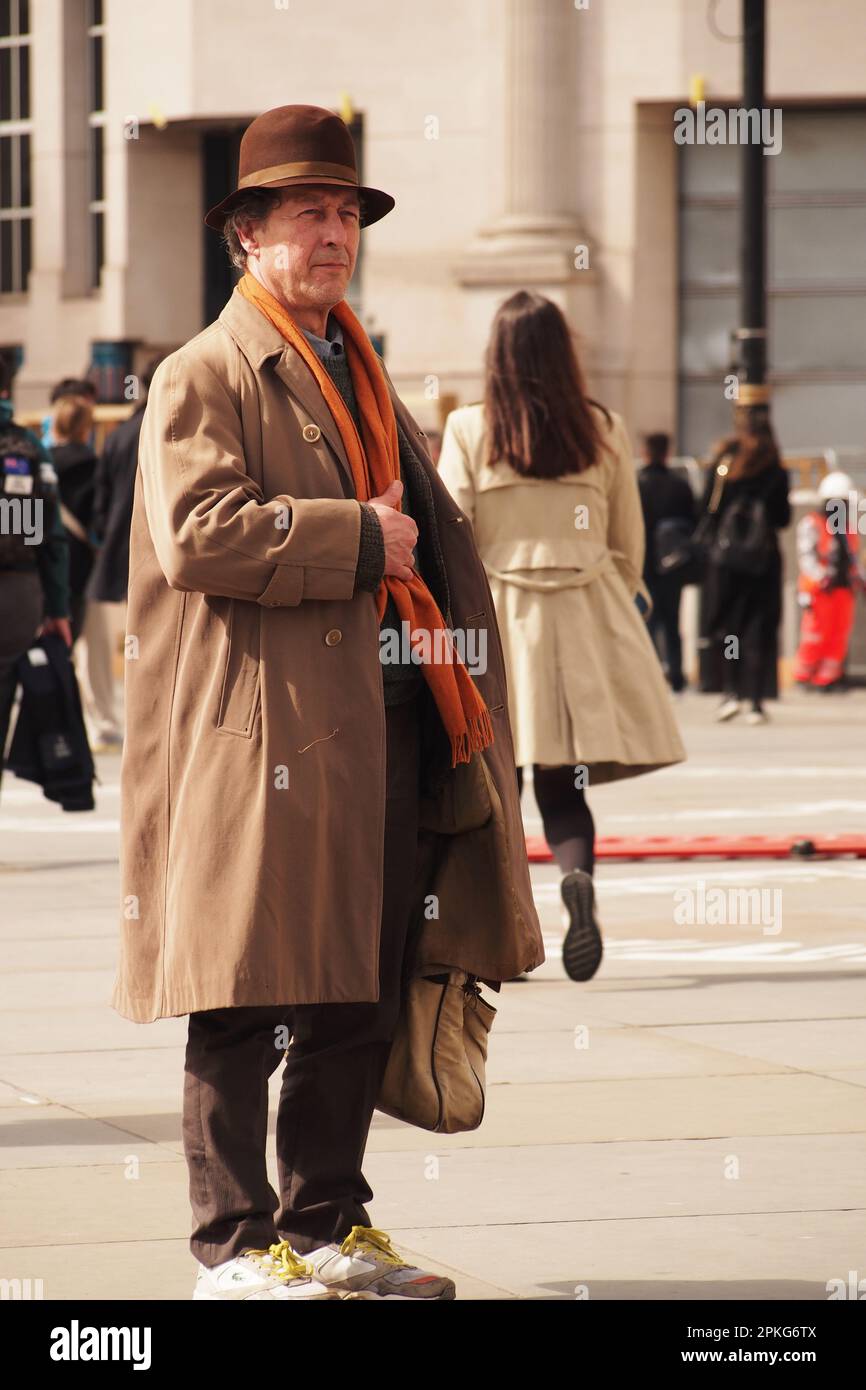 An older man 60+ standing with long coat, large bag, orange scarf, brown hat standing in Trafalgar Square, London watching and listening to a busker Stock Photo