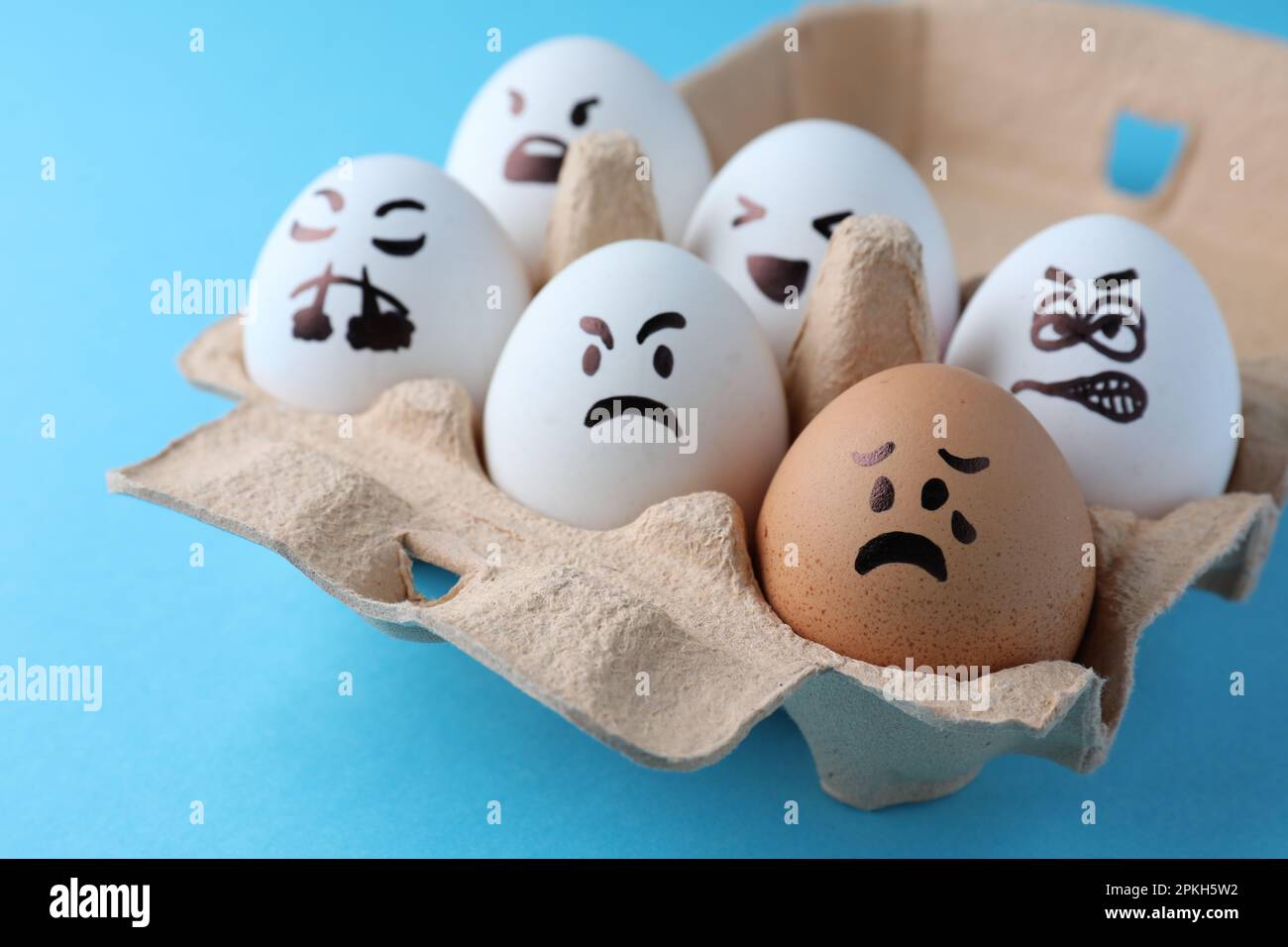 Brown egg with upset face among aggressively disposed white ones in carton box on turquoise background, closeup. Bullying concept Stock Photo