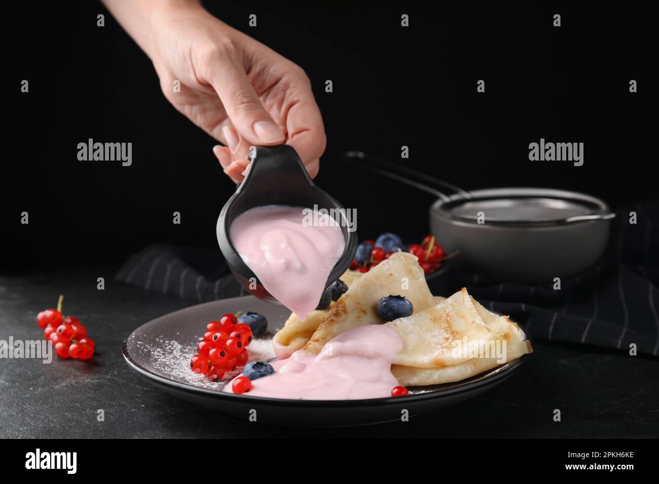 Woman pouring natural yogurt onto crepes with blueberries and red currants at table, closeup Stock Photo