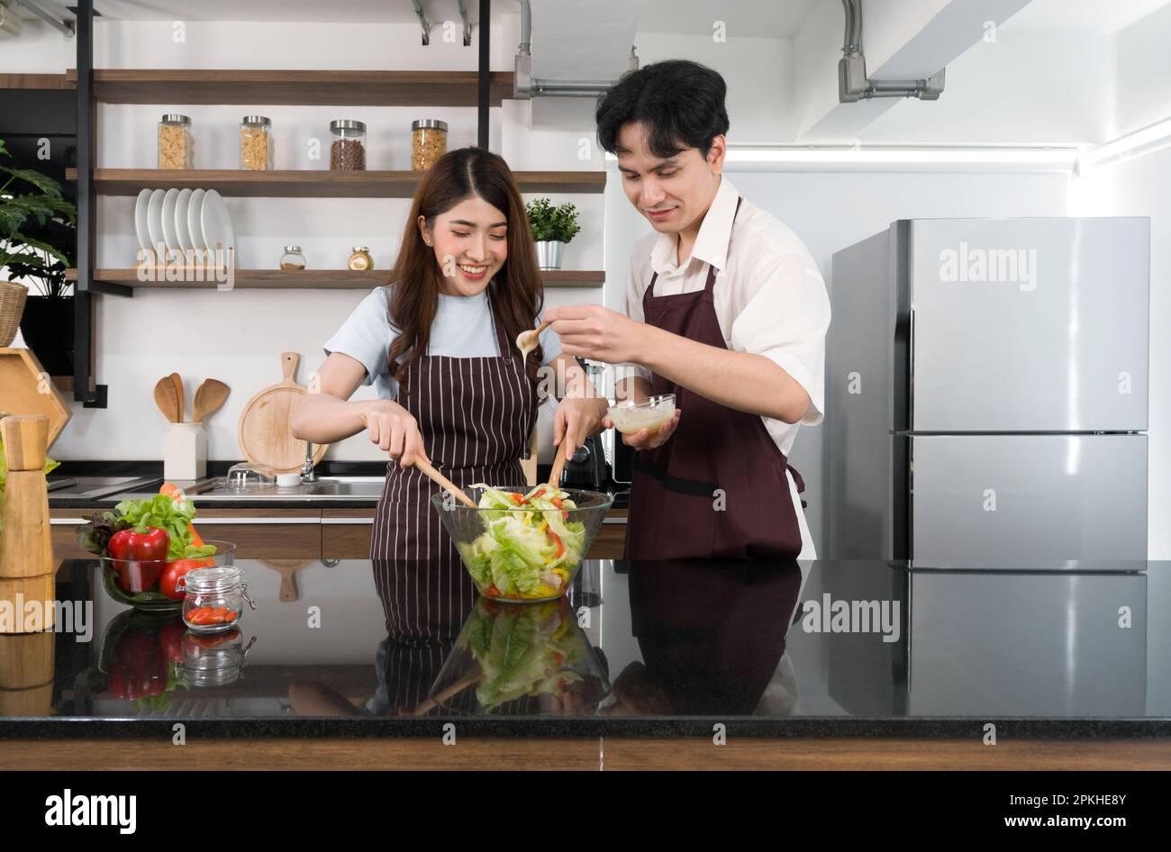 Young asian man dressed in an apron putting salad dressing in a salad bowl  while his girlfriend mix the vegetables together with a wooden ladle. Tomat  Stock Photo - Alamy