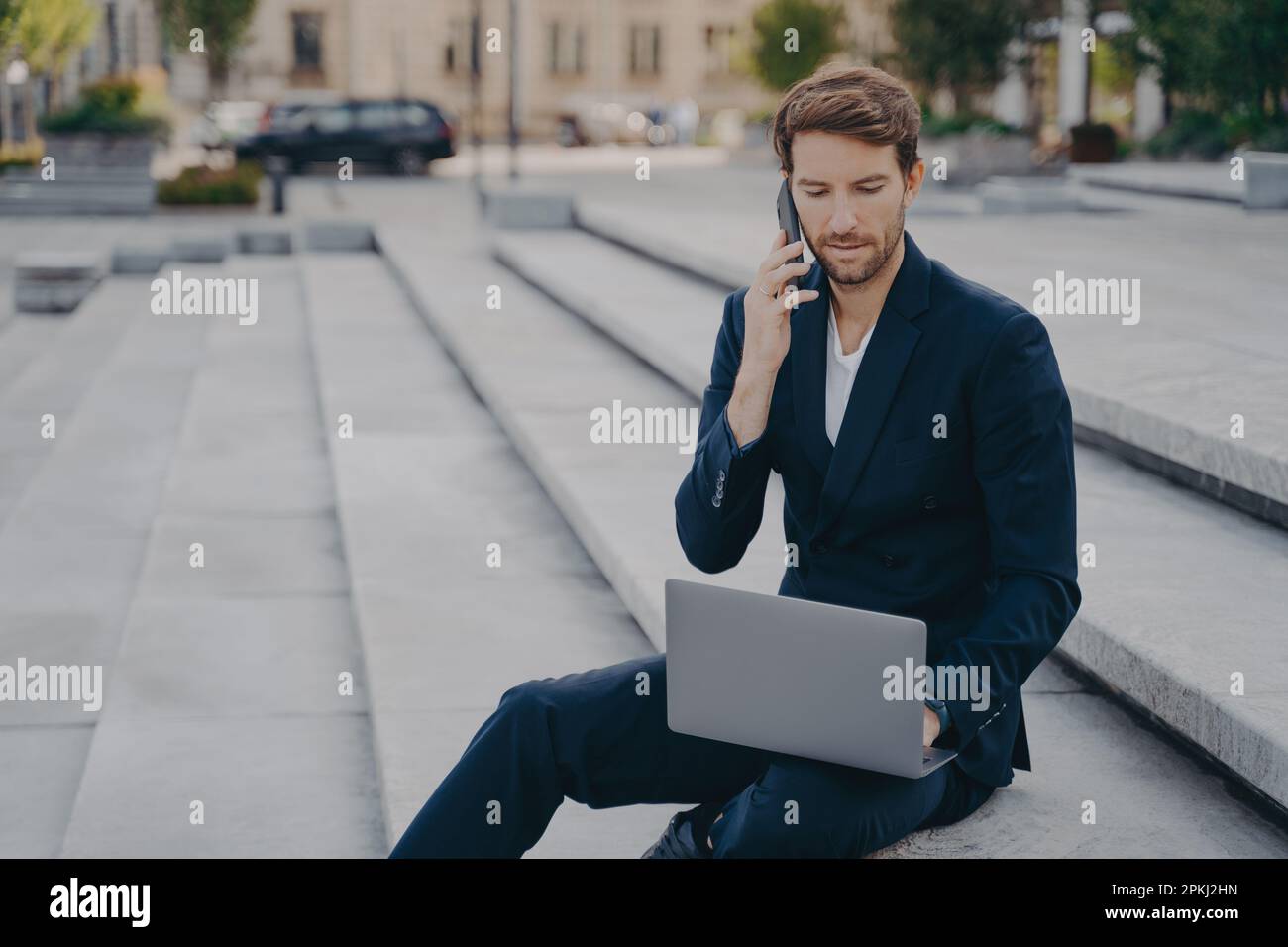Man executive worker talks on cellphone with business partner uses modern laptop computer for making startup project dressed in formal suit sits on st Stock Photo