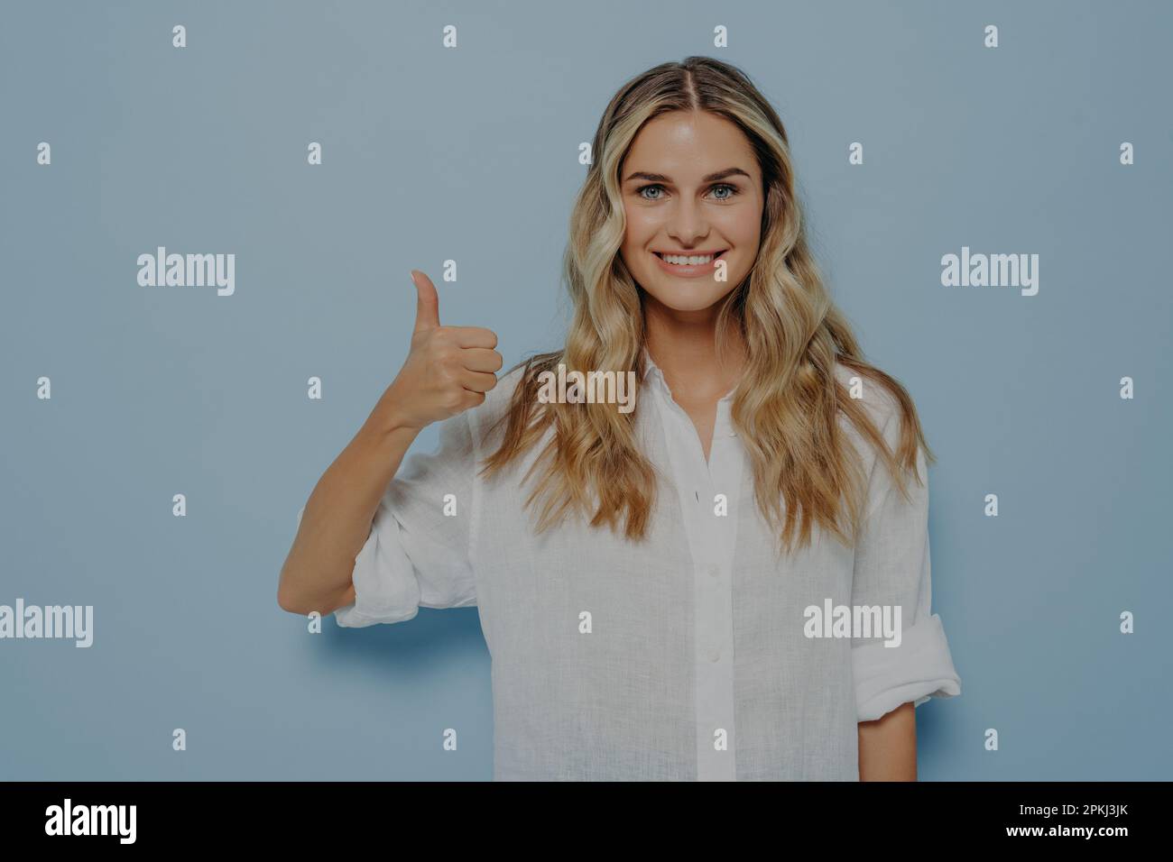 Cheerful blonde girl in white shirt showing thumbs up gesture with her hand while smiling, admitting that she liked that, standing alone next to blue Stock Photo