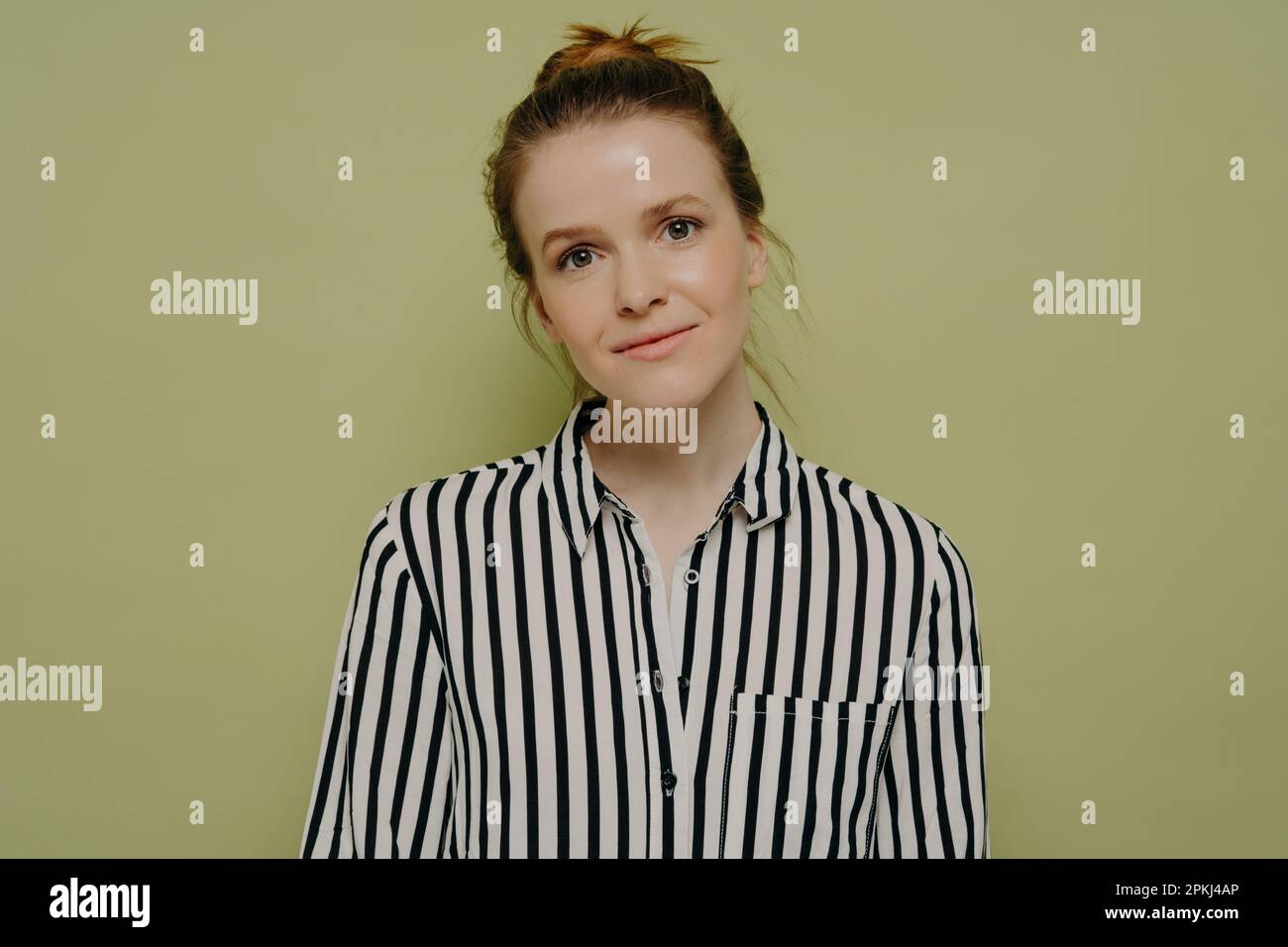 Portrait of calm young european woman with hair in bun wearing black and white striped shirt looking at camera with tilted head and slight smile while Stock Photo