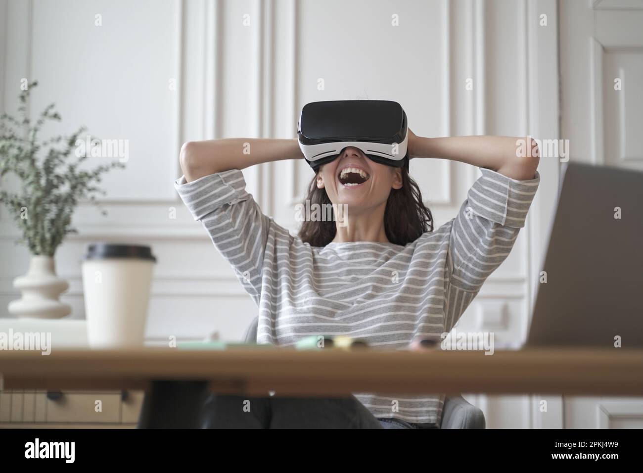 Amazed woman office employee in VR glasses laughing with joy leaning back in chair with hands behind head while remotely working from home office, che Stock Photo