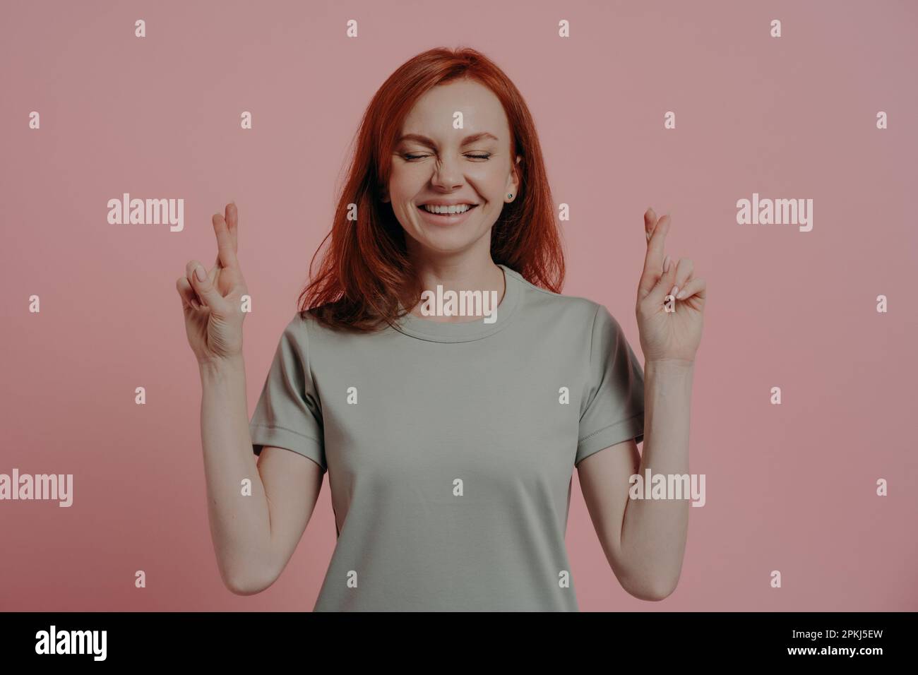 Studio shot of hopeful red-haired female student with closed eyes in casual wear crossing fingers with superstitious facial expression, asking for goo Stock Photo