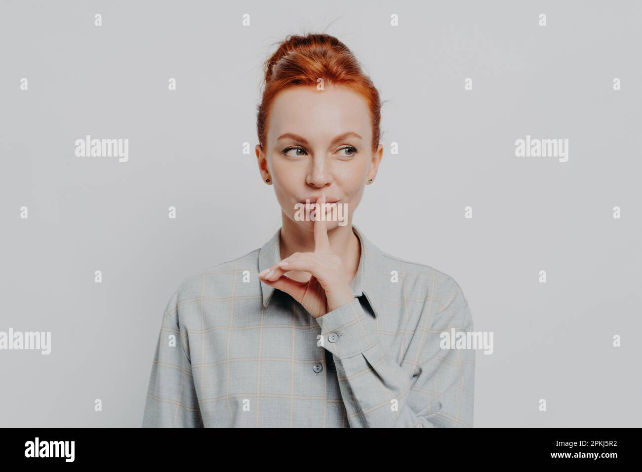 Keep it in secret. Young ginger woman with hair in bun showing silence sign, keeping finger on lips and looking aside at copy space while posing isola Stock Photo