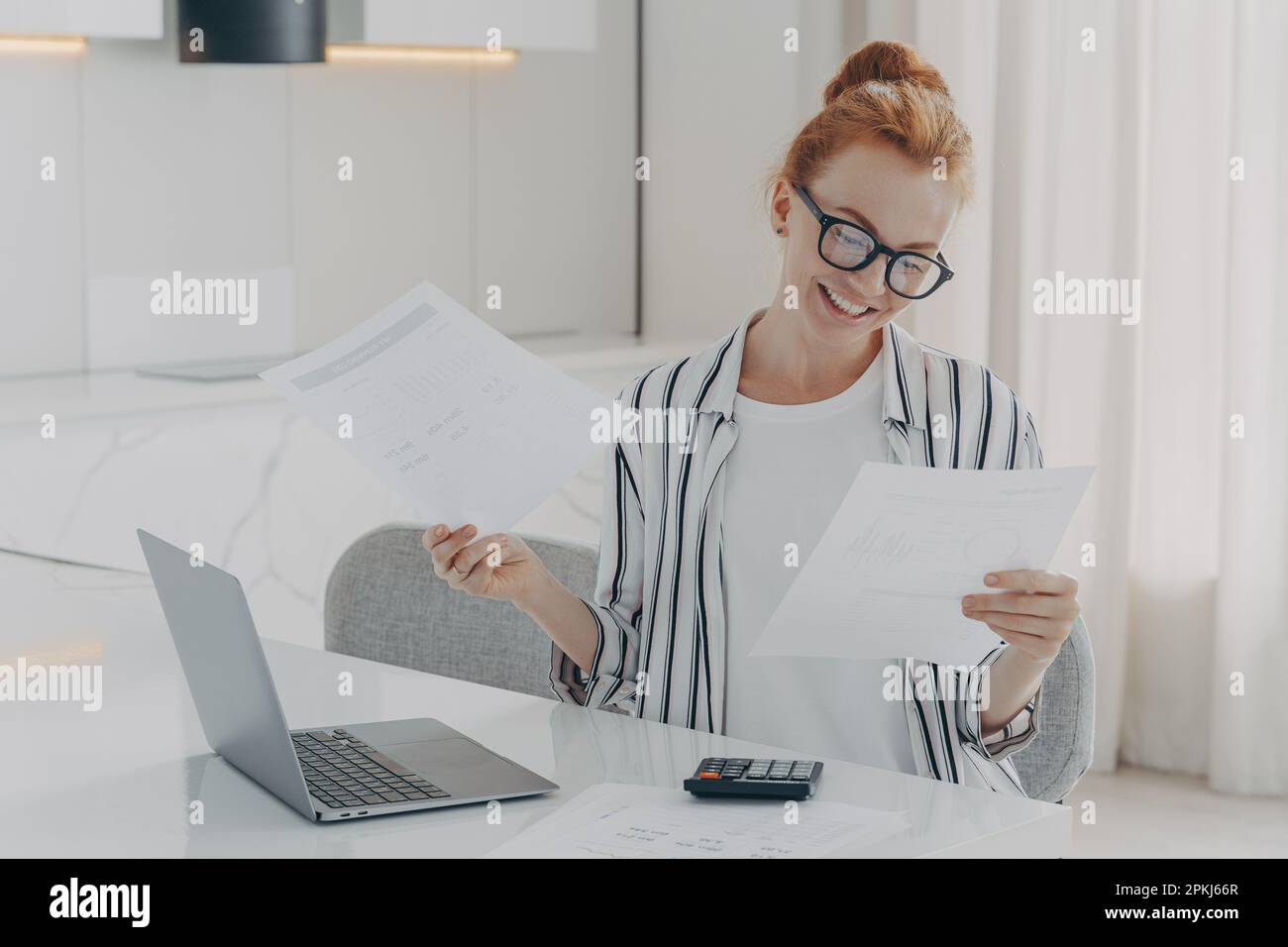 Casually dressed excited redhead woman sitting at table at home, feeling happy after checking domestic bills, holding holding documents with bank noti Stock Photo