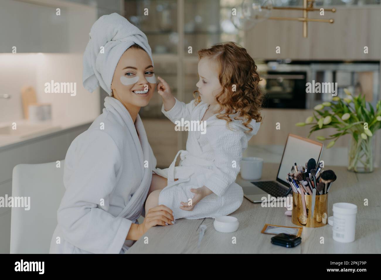 Happy young mother teacher her little daughter to do makeup. Culry small girl applies beauty patches under eyes to mom use beauty products naturl cosm Stock Photo