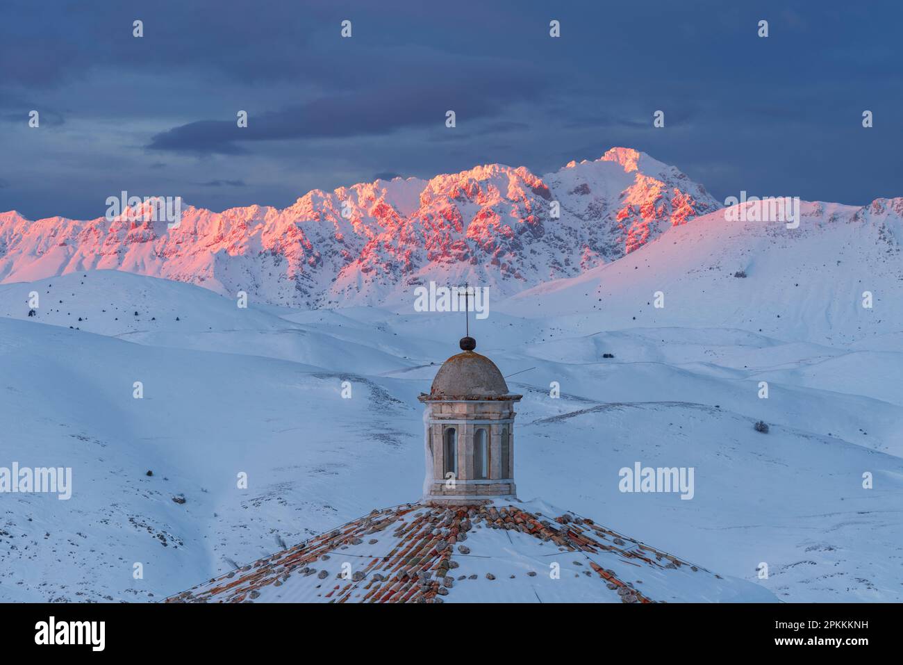 Close-up view of the roof of the church of Santa Maria della Pieta with snow covered mountains of Gran Sasso lit at sunset, Rocca Calascio Stock Photo