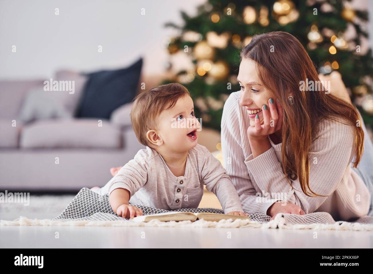 With book that is on the ground. Mother with her little daughter is indoors at home together. Stock Photo