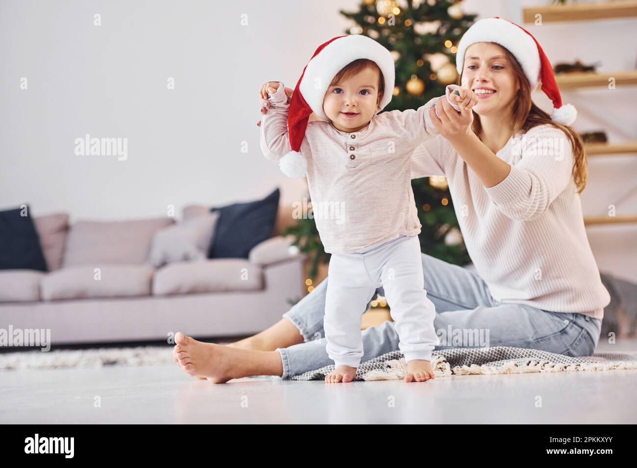 In Santa hats. Mother with her little daughter is indoors at home together. Stock Photo