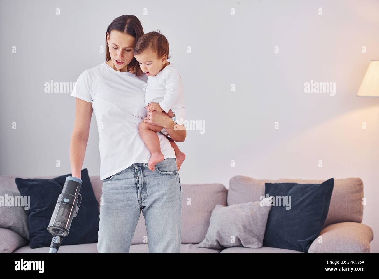 Woman uses vacuum cleaner. Mother with her little daughter is indoors at home together. Stock Photo