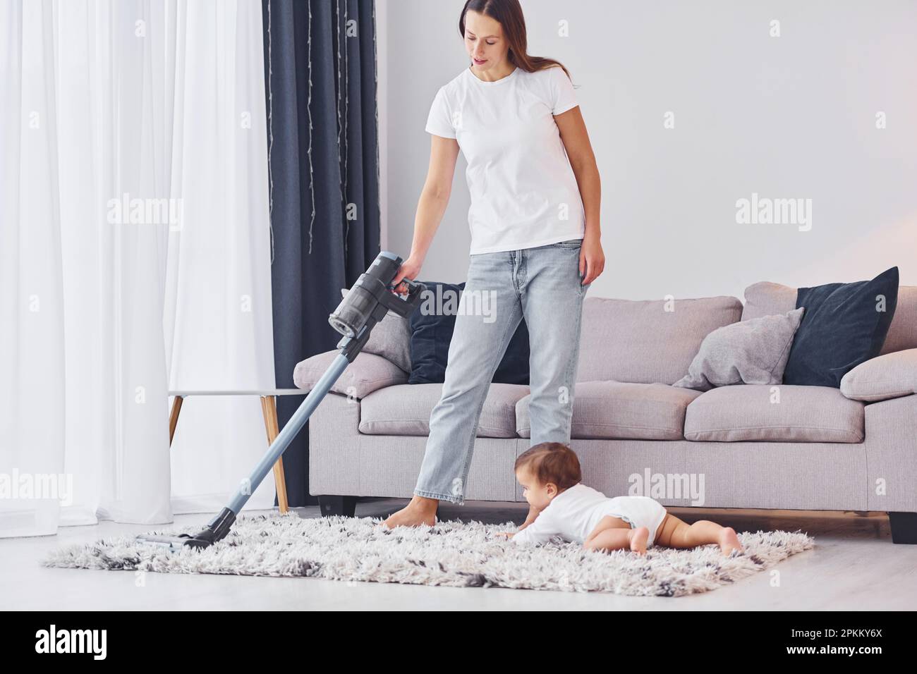 Using of the vacuum cleaner. Mother with her little daughter is indoors at home together. Stock Photo