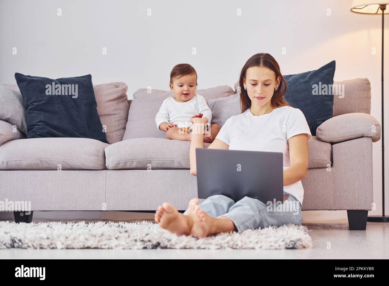 Woman working by using laptop. Mother with her little daughter is indoors at home together. Stock Photo