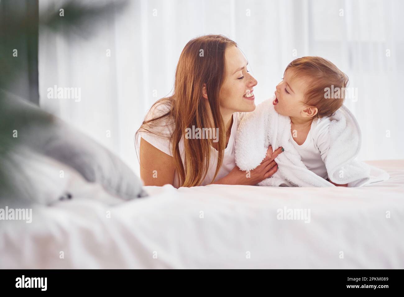Laying down on the bed. Mother with her little daughter is indoors at home together. Stock Photo
