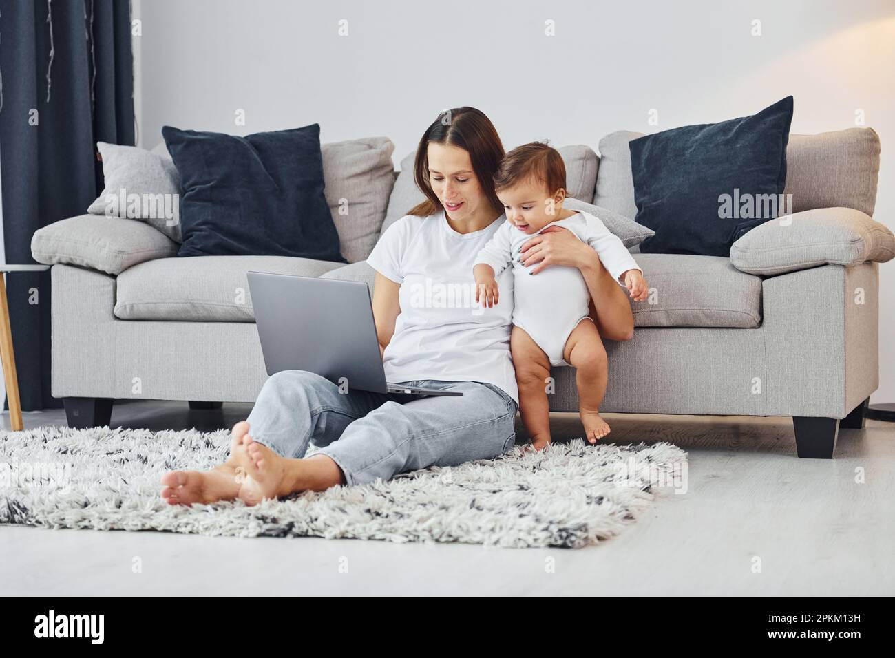 Silver colored laptop. Mother with her little daughter is indoors at home together. Stock Photo