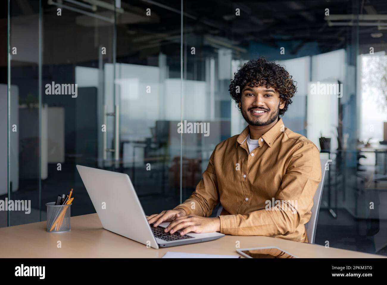 Portrait of young successful Indian man at workplace inside office, businessman smiling and looking at camera, man at work using laptop, programmer with curly hair coding software. Stock Photo