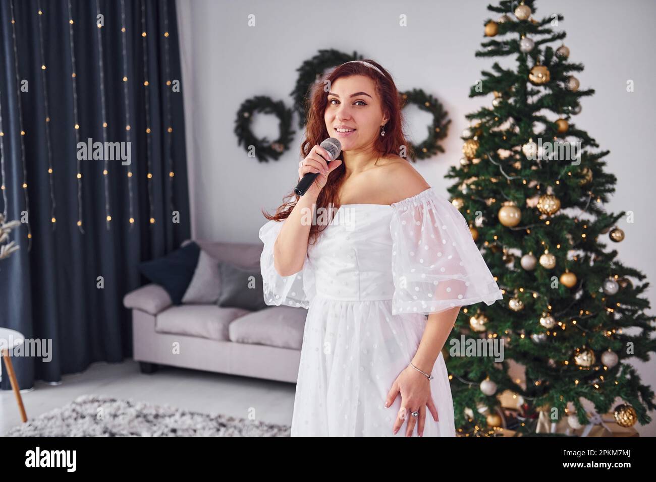Standing in the christmas decorated domestic room. Woman in white dress and with microphone in hands is singing in the . Stock Photo