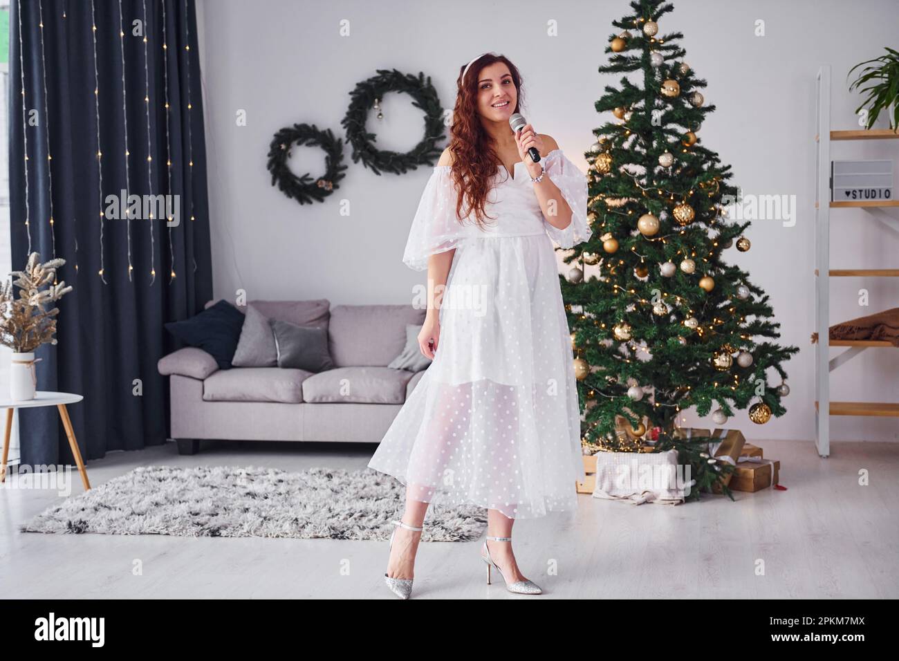 Woman in white dress and with microphone in hands is singing in the christmas decorated domestic room. Stock Photo