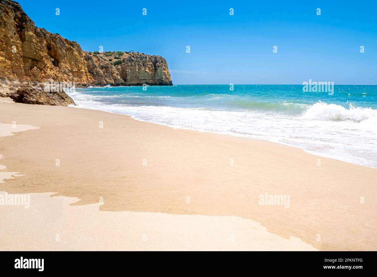A picturesque and secluded Algarve beach, Praia do Canavial, boasts fine sand and blue waters with a small wave breaking under the bright sun. Stock Photo