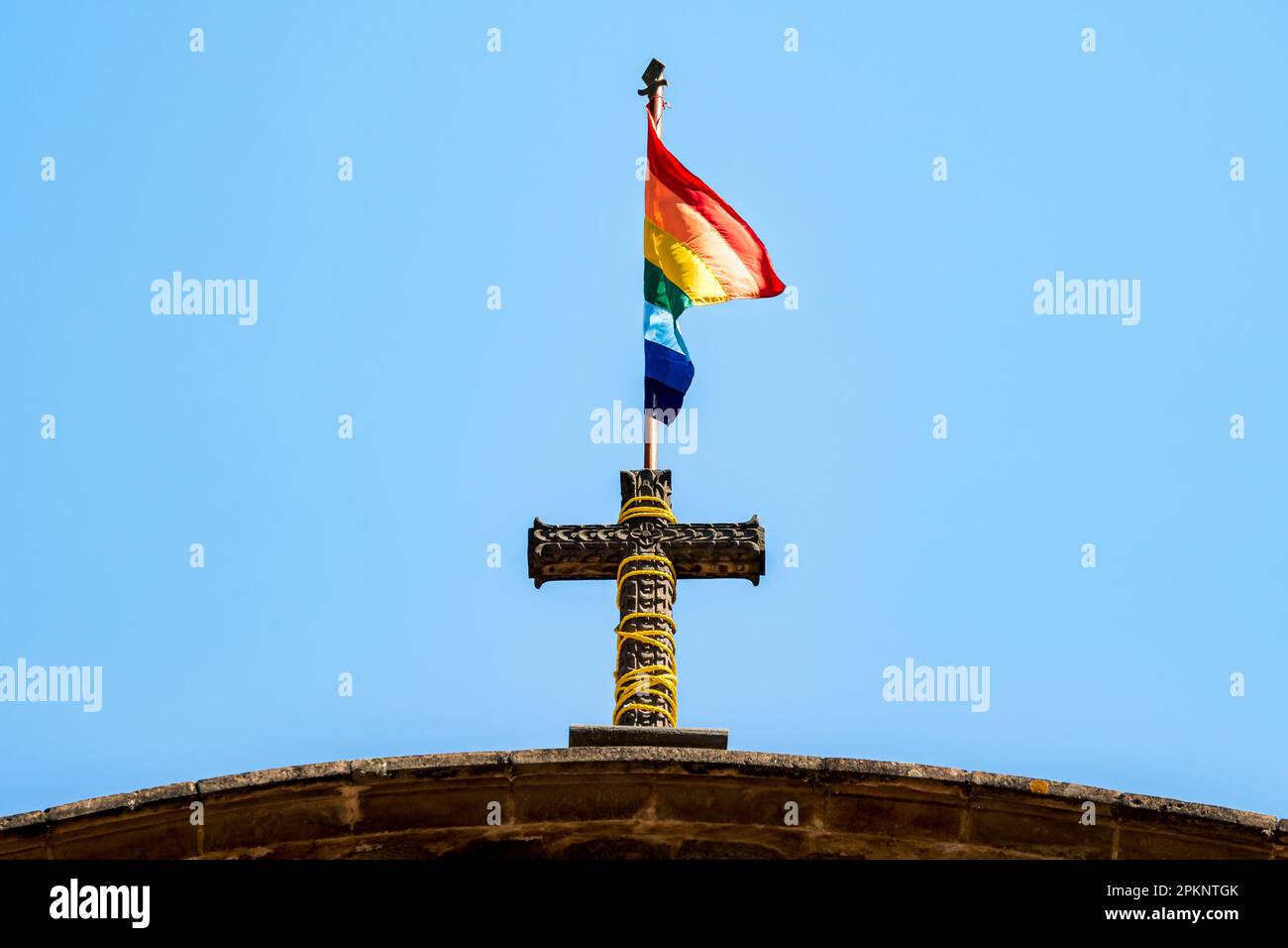 The rainbow flag of Cusco is proudly attached to a religious cross outside of a church, symbolizing the importance of pride, faith and unity. Stock Photo