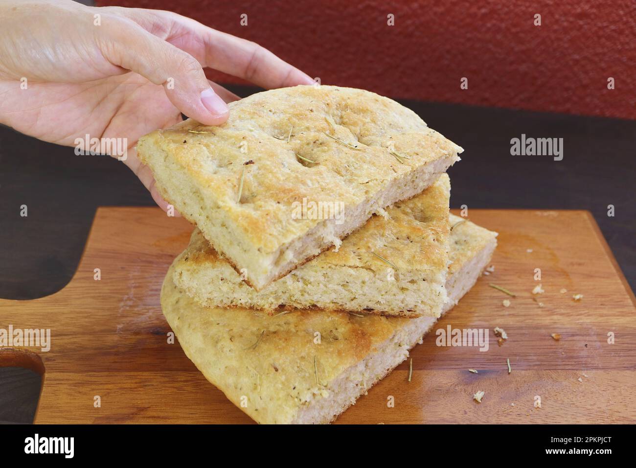 Closeup Hand Picking a Slice of Tasty Rosemary Focaccia Bread Piled Up on Wooden Breadboard Stock Photo