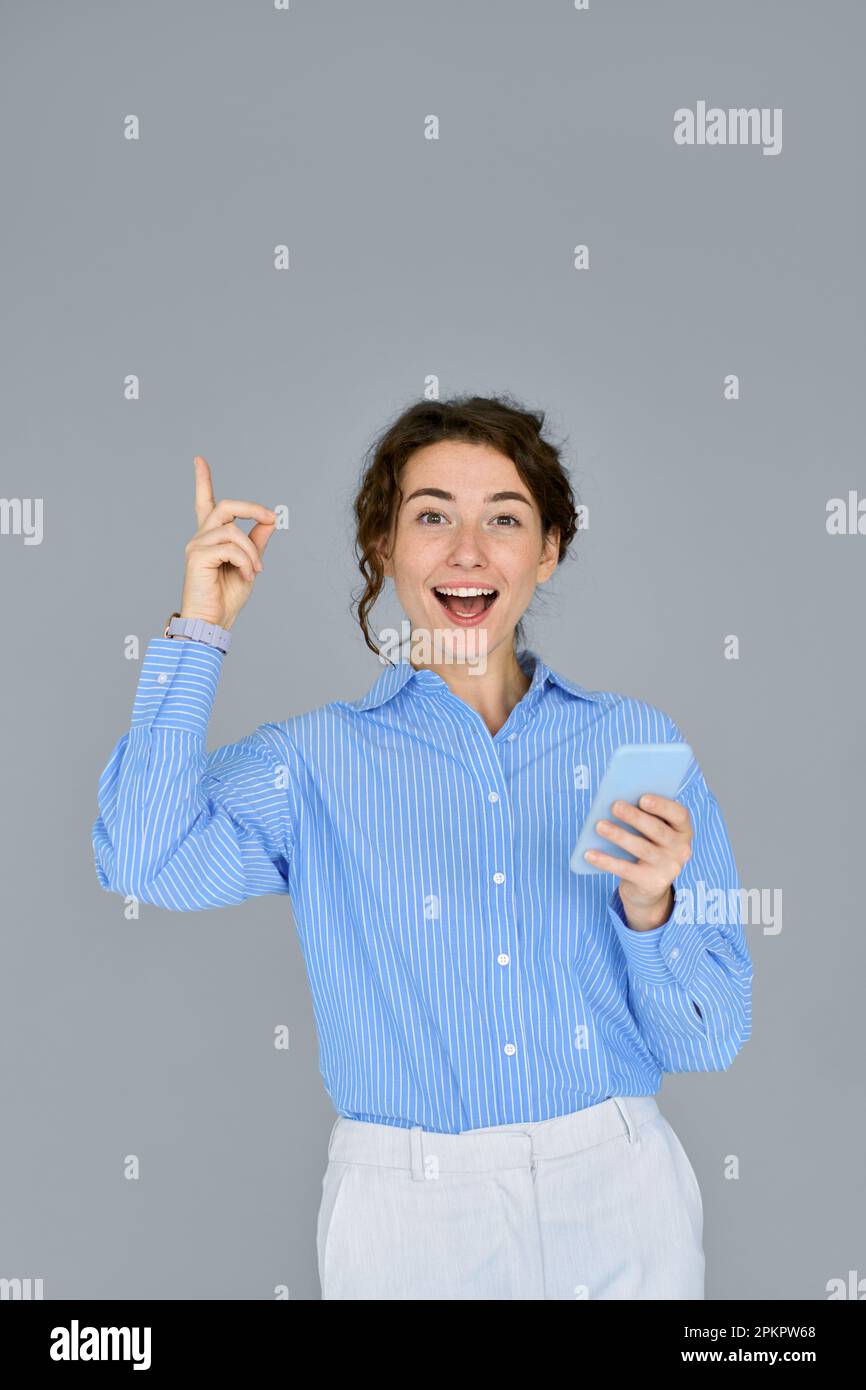 Young happy surprised business woman using phone pointing up isolated on grey. Stock Photo