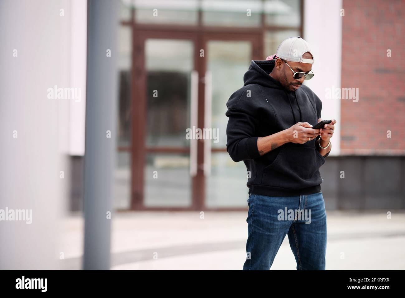 African American black man uses mobile phone, stands on city street, wears hoodie, baseball cap and sunglasses. Portrait Stylish male model looks at h Stock Photo