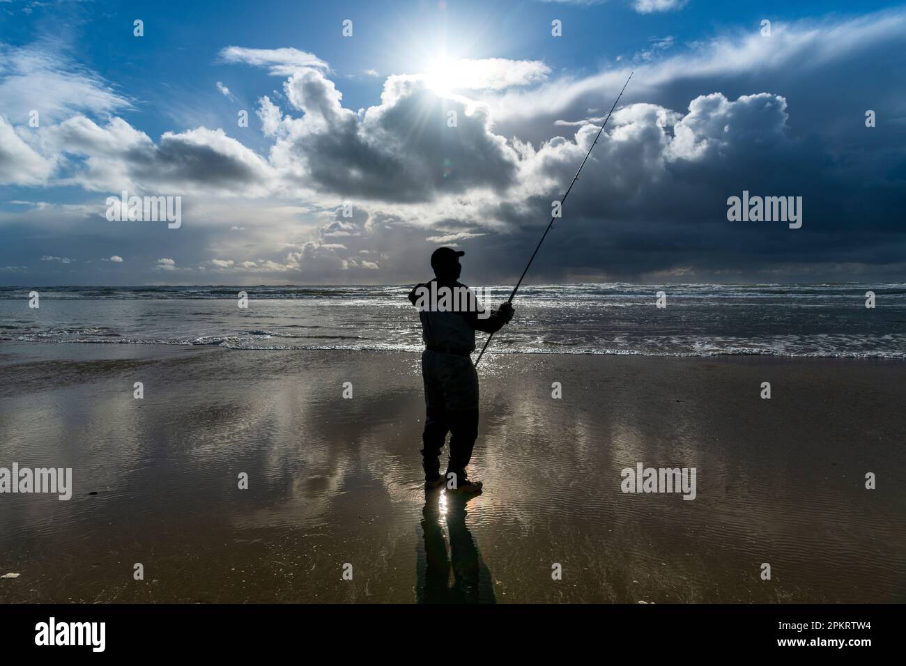 A solo fisherman in wet suit standing at ocean shore during low tide,  reflecting in the wet shore sand and backlit by the sunset light Stock  Photo - Alamy