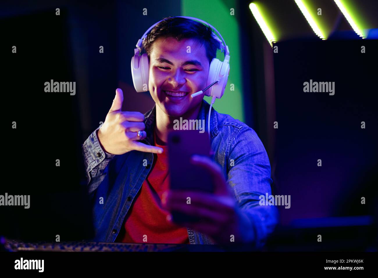 Smiling Gen Z gamer connecting with streamers and players in an online gaming community using a smartphone. With a headset on, he engages in a live vi Stock Photo