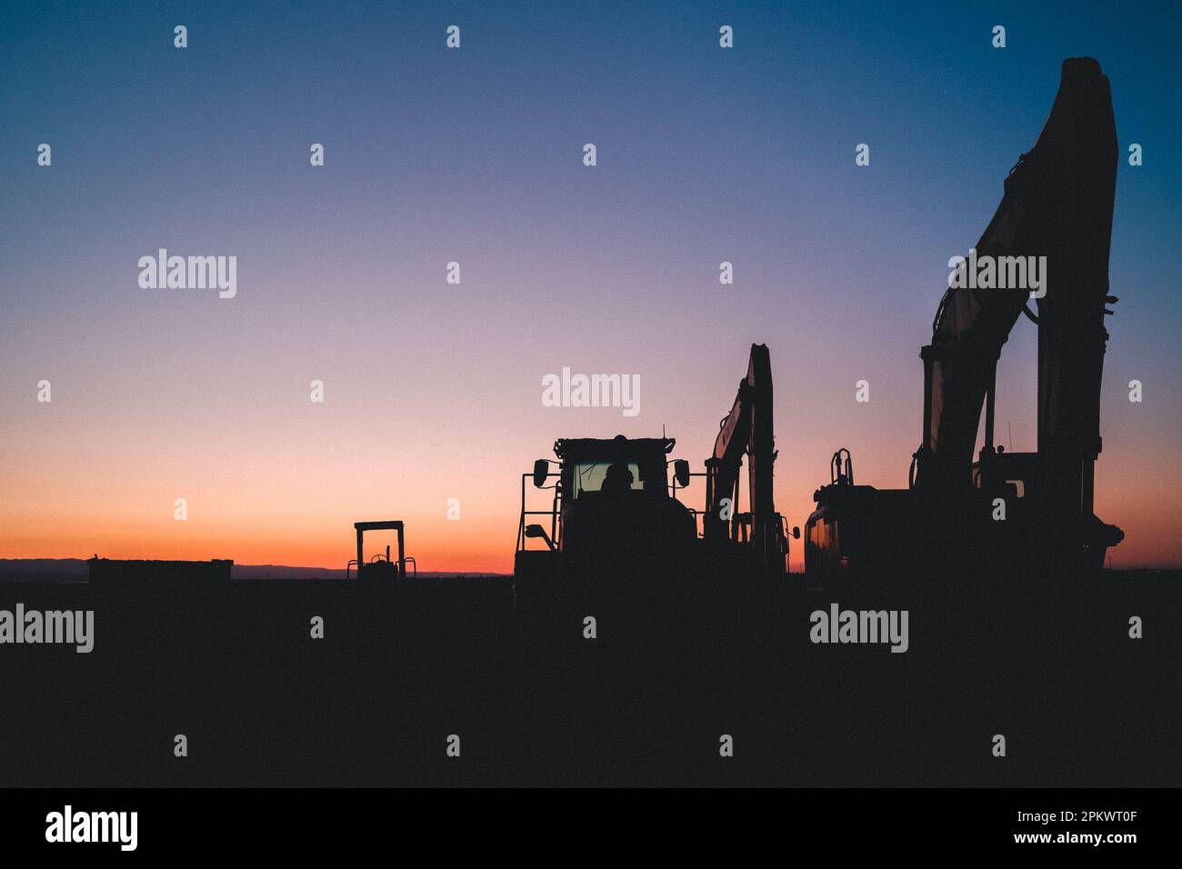 The silhouettes of two industrial machines standing in a field at dusk, illuminated by the setting sun. Stock Photo