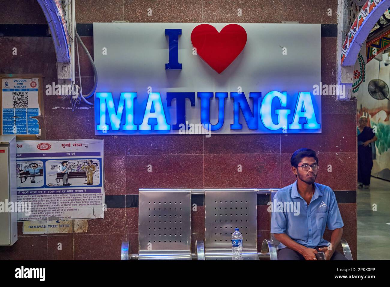 A male passenger sits on a bench at Matunga railway station (Central Railways), Mumbai, India, under an illuminated neon sign saying 'I Love Matunga' Stock Photo
