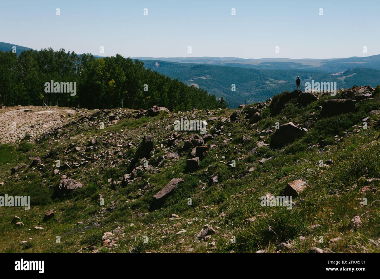 Boy standing on rocky hill looking down at valley in mountain hike Stock Photo