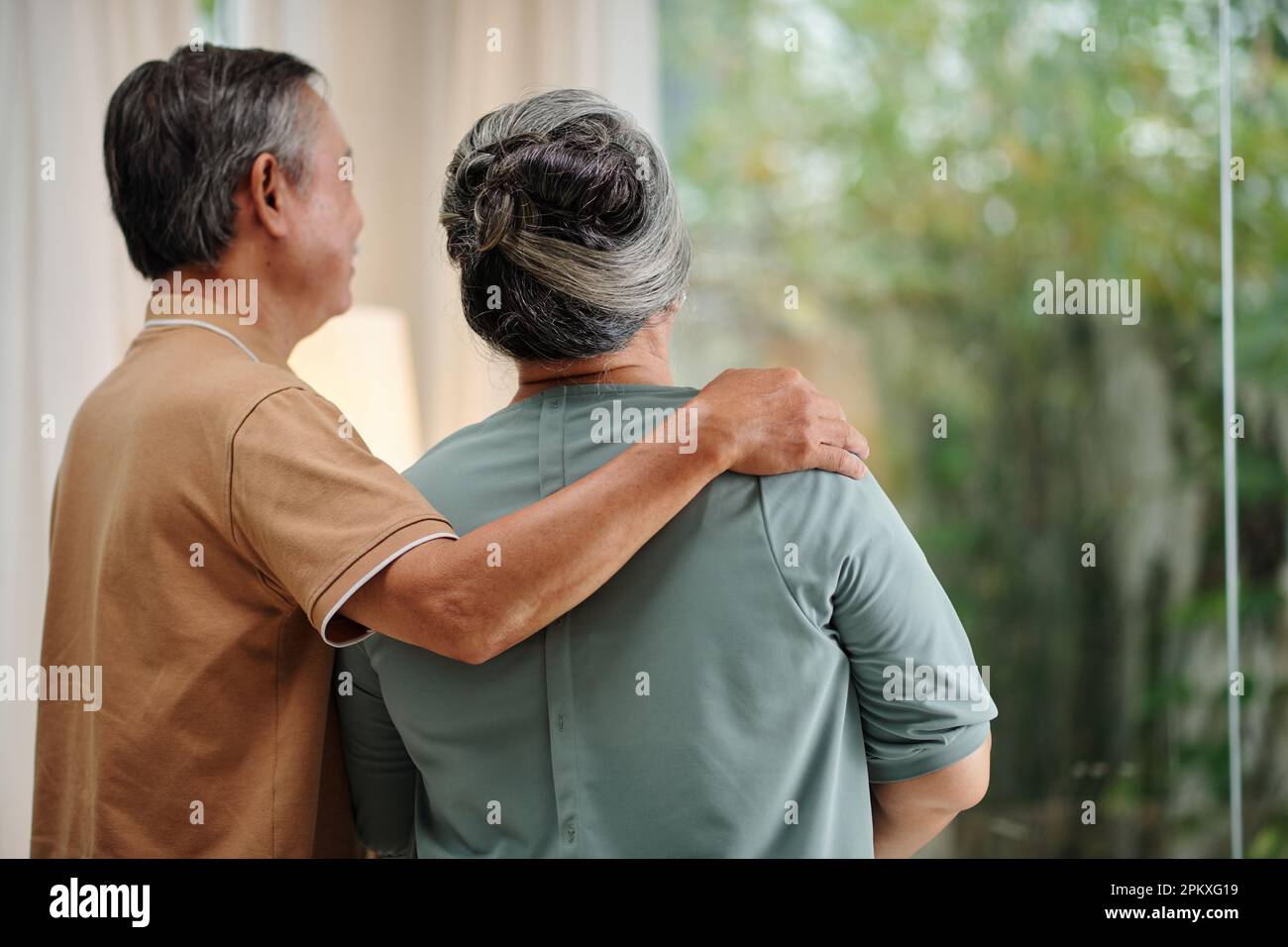 Hugging couple looking outside through big window Stock Photo - Alamy