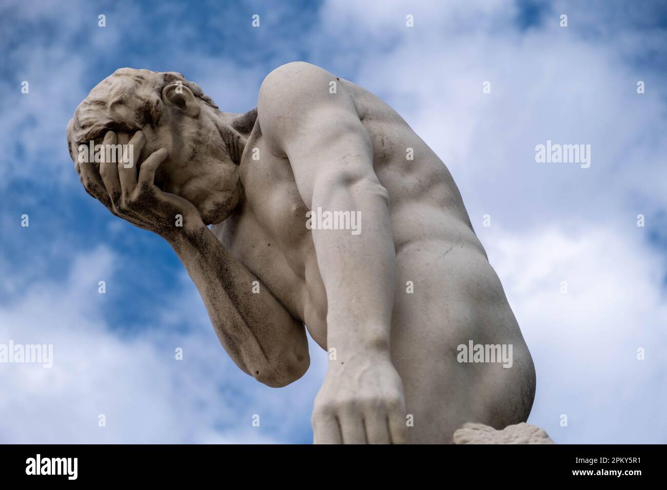 White Stone Statue of Muscular Man with Hand on Face, Against Blue Sky and Clouds Stock Photo