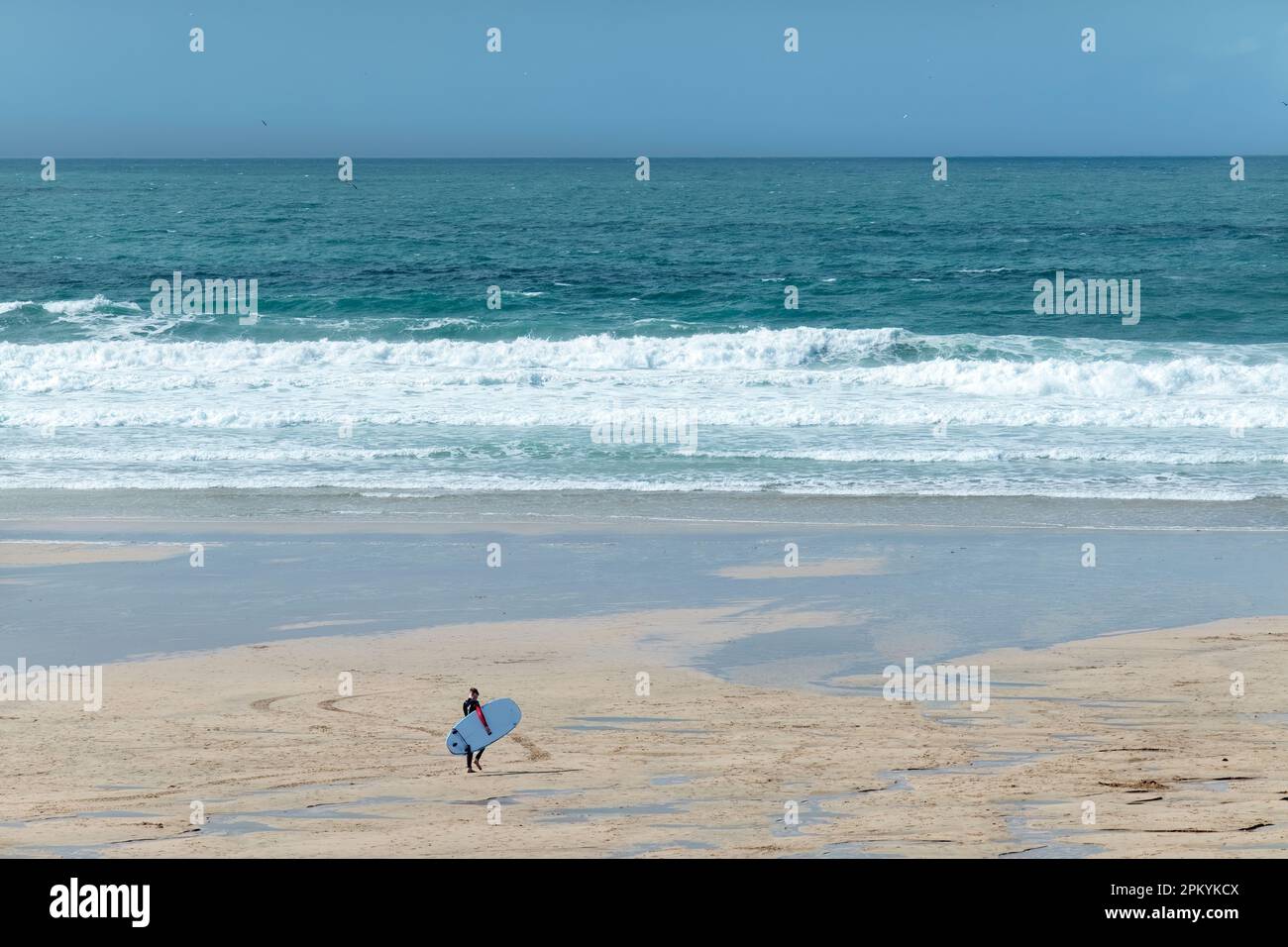 A lone male surfer walks out towards the sea on Fistral Beach, Newquay Cornwall. He's carrying his board as he crosses the beach towards the waves Stock Photo