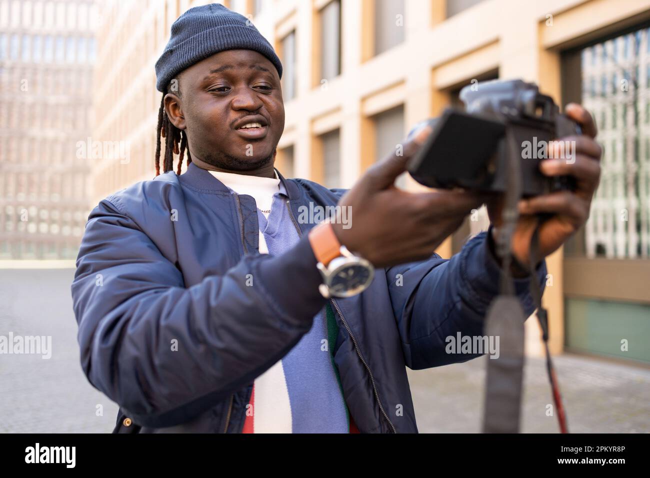 Happy black young male in trendy clothes with braids standing outside and looking at camera while taking self photo on professional camera Stock Photo