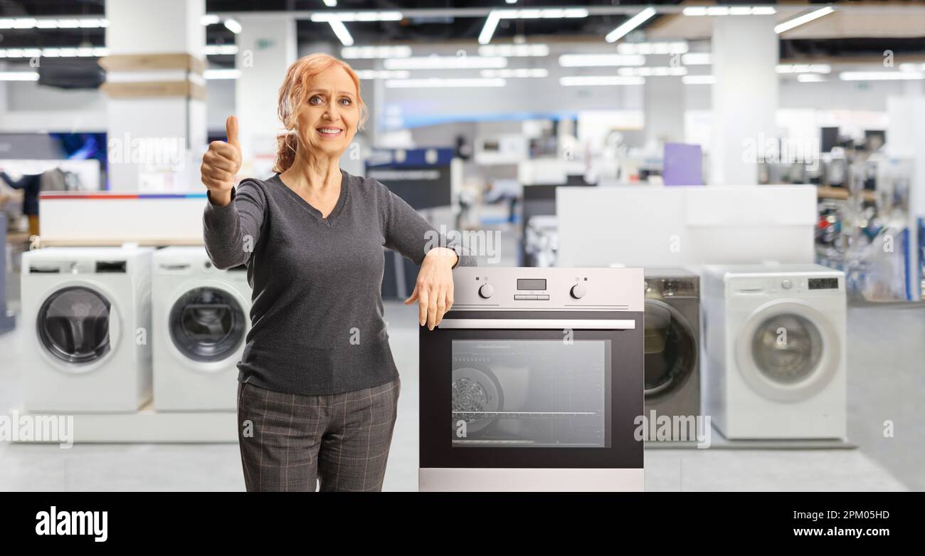 Happy mature woman posing with home applances and gesturing thumbs up inside a store Stock Photo