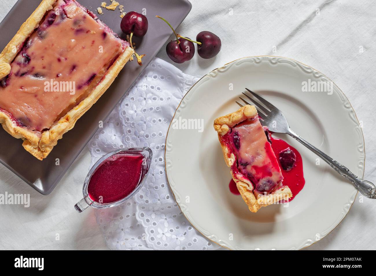 A slice of a ricotta cheese and cherries homemade cake. At side: the cake, some cherries and a small glass gravy boat with syrup. Stock Photo